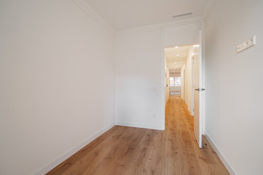 A bright, empty hallway with white walls and wooden flooring. The space is well-lit, leading to an open area in the background.