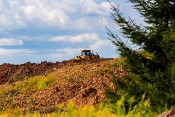 Yellow bulldozer pushing dirt on a construction site profile view of heavy industrial machinery working on a mound under a bright blue sky with fluffy clouds near pine trees © PhotoChur