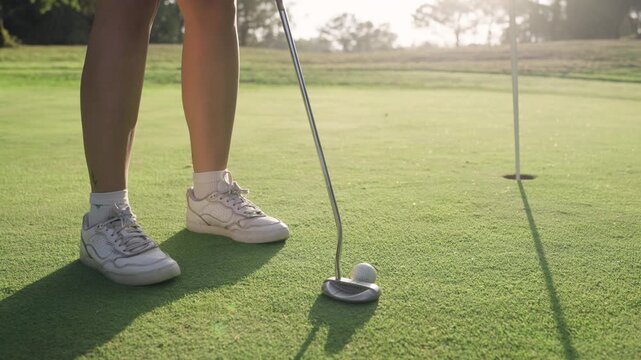 Low angle shot of a woman's legs and putter as she successfully makes a putt. The golf ball rolls smoothly across the green into the hole