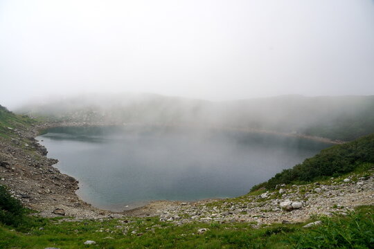 日本の山岳地帯に位置する火口湖の風景。霧が立ち込める神秘的な雰囲気の中、岩場に囲まれた青い湖面が静かに広がっている夏の山の情景。