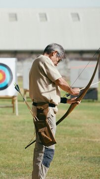 Elderly man with gray hair concentrating as he prepares his longbow for shooting at an outdoor archery range. Active senior enjoying his hobby. Vertical