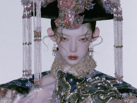 Ornate ancient‑style BJD doll wears elaborate embroidered royal costume, pearl tassel headdress against plain white background with delicate facial makeup.