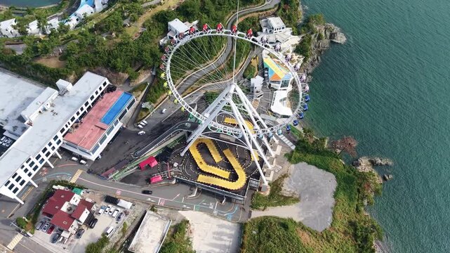 Close Aerial Drone View of Massive White Ferris Wheel Structure at Yeosu Art Land Coastal Amusement Attraction, Detailed Observation Wheel Architecture Surrounded by Rocky Shore and Blue Ocean in Sout