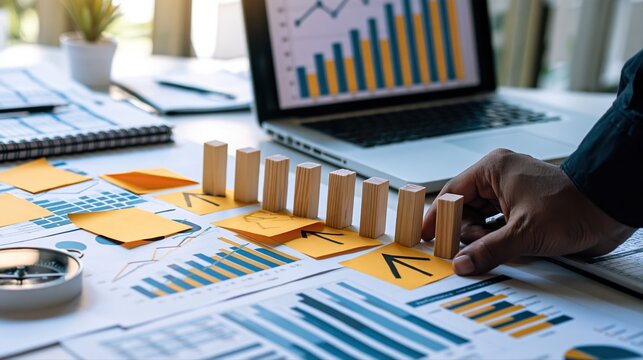 A business strategy desk with charts, a laptop, and wooden milestone blocks. A hand arranges progress blocks, symbolizing planning and forward growth.