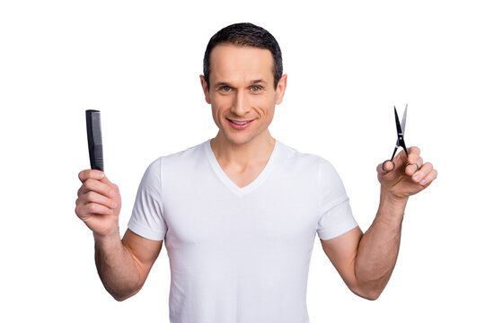 Portrait of experienced, neat, brunet hair expert, stylist having scissors and comb in arms, looking at camera, wearing white t-shirt, standing over grey background, let's make a trendy hairstyle