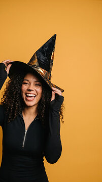 Curly-haired woman smiles joyfully in a witch hat. She wears a black costume against a warm orange background. Her playful pose exudes Halloween spirit and festive cheer