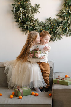 Loving sister embracing her little brother holding a Christmas gift in a decorated home livingroom with evergreen wreath, presents, and tangerines during holiday celebration.