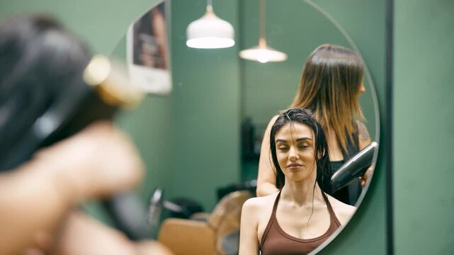 Mirror reflection of a female hairdresser using a hairdryer on a client's wet hair. Professional hair care and styling in a beauty parlor