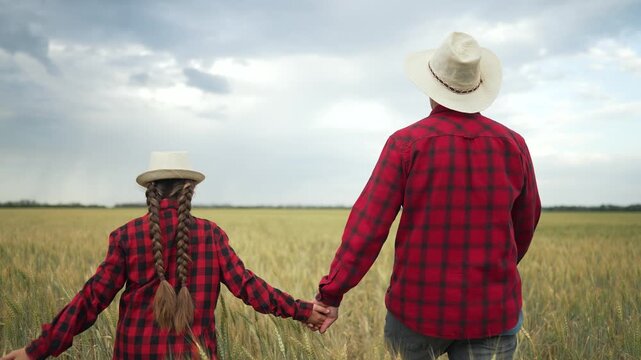 Father and daughter walking through field. Family walk together nature. Matching red plaid shirts and straw hats. Farmer family bonding moment. Scenic countryside family walk. Happy family walk field.