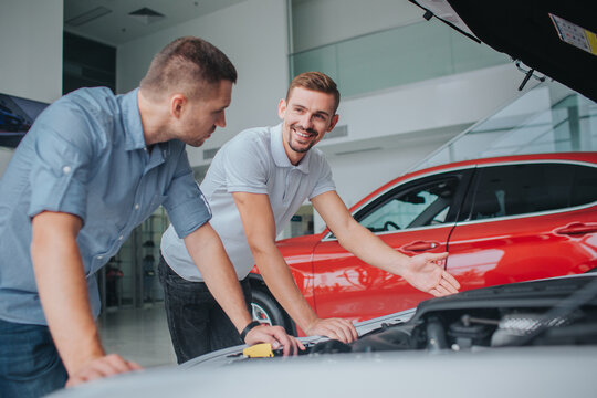 Seller in white sirt stands at opened body of car and point on it. He looks at guy and smiles. Man in grey shirt leans on car and looks at seller. He choose car.