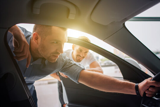 Handsome and confident customer stands and leans to car. He holds steering wheel with one hand. Guy is attentive to details. Manager stands behind him and lean to car. He is smiling and watching.