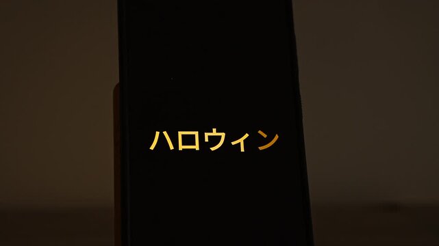 Halloween written in Japanese katakana, ハロウィン, glows in warm orange on a smartphone standing in a dark room. A spooky season greeting for Japanese Halloween events, October 31 parties and localized h