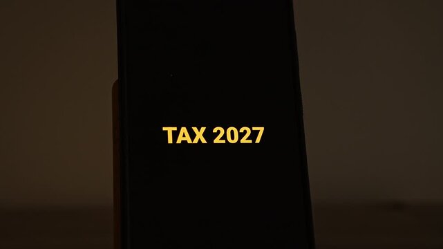 TAX 2027 types itself out in orange gold letters on a smartphone standing alone in a dark, warm room. A clean title for tax year 2027, new tax rules, filing deadline reminders and small business acco