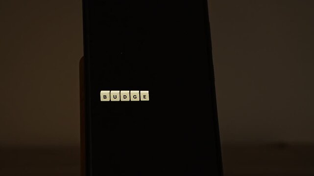 Cream letter cubes spell BUDGET 2026 on a smartphone standing in a dim room, and the last cube flips to turn the year into 2027. It shows budget rollover, next year financial plans, company budgeting
