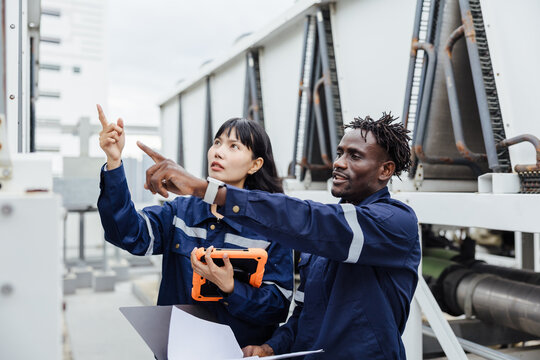 Two technicians in protective uniforms inspect large rooftop HVAC units, pointing and discussing maintenance tasks with a tablet and clipboard at an industrial facility.