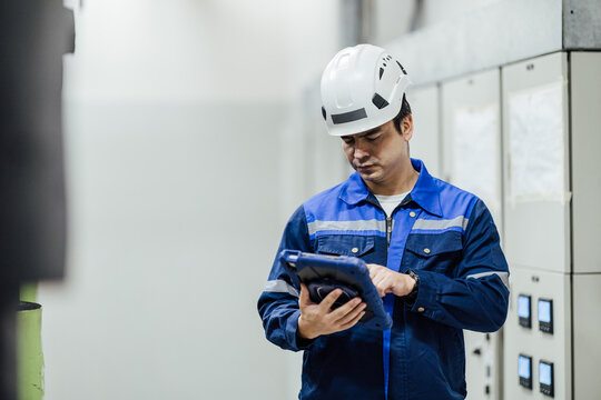 Industrial technician in hard hat using a tablet to inspect electrical control panels inside a factory, emphasizing safety, maintenance, and digital operations.