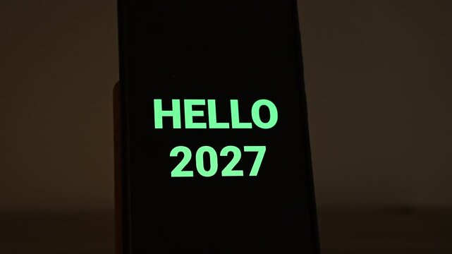 HELLO 2027 appears in fresh mint green, its last digits fading in, on a smartphone standing in a dim, warm room. A positive opener for the first day of the year, new goals, welcome messages and Janua