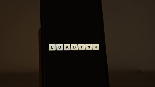 Small cream tiles spell LOADING on a smartphone in a dim room, then rearrange themselves into 2027. A neat reveal for the coming year, new year loading concepts, anticipation and 2027 launch teasers.