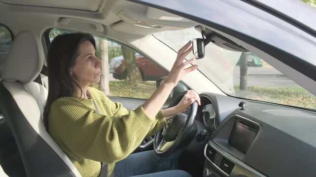 Latin American woman adjusts rearview mirror inside automobile carefully preparing for journey while sitting behind steering wheel during bright daytime commute preparations within vehicle