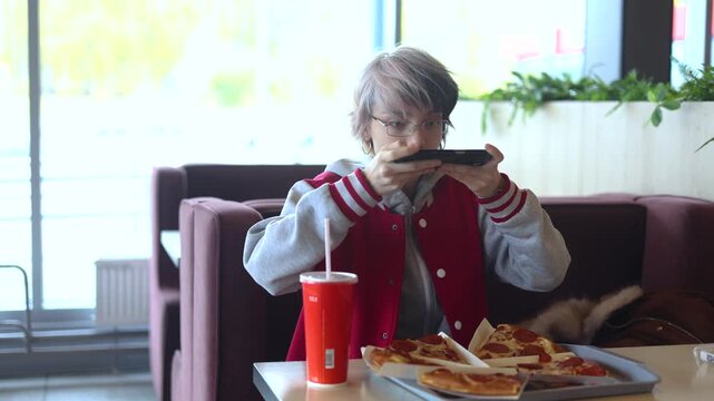 The teenage girl is sitting in a cafe and taking a photo of her pizza with her smartphone. She looks at the photo, smiles, and sends the photo to her friends on social media. Slow motion 4K video.