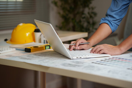 An architect reviews residential floor plans and color palettes while working on a laptop with construction tools and a miniature house model.