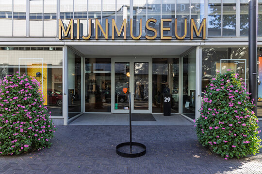 Heerlen, Netherlands - October 04, 2026: Frontal view of Nederlands Mijnmuseum glass entrance with yellow neon sign and decorative flower bushes.