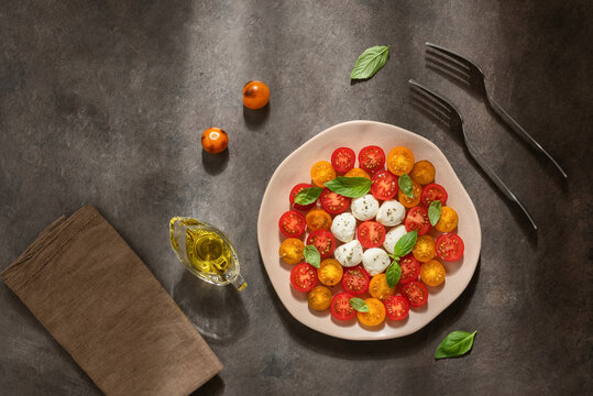 Caprese salad with mozzarella, colorful cherry tomatoes and basil leaves in a plate on a dark rustic background in sunlight, top view, flat lay