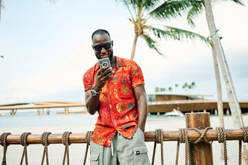 A man stands on a sunny tropical beach checking his smartphone while wearing a red patterned shirt and sunglasses. Casual vacation vibes and modern lifestyle combine as he texts or scrolls near a mari © Lumos sp