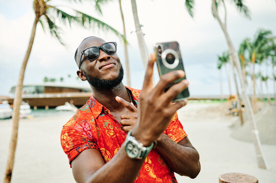 A smiling man takes a selfie on a tropical beach lined with palm trees while wearing sunglasses and a bright patterned shirt. He holds a smartphone, enjoying a relaxed vacation moment at a sunny resor