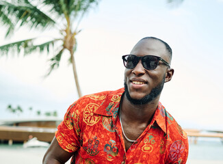 A smiling man wearing sunglasses and a bright red Hawaiian shirt enjoys a sunny tropical beach setting with palm trees and calm ocean waters. Casual and relaxed, the portrait conveys vacation, leisure © Lumos sp