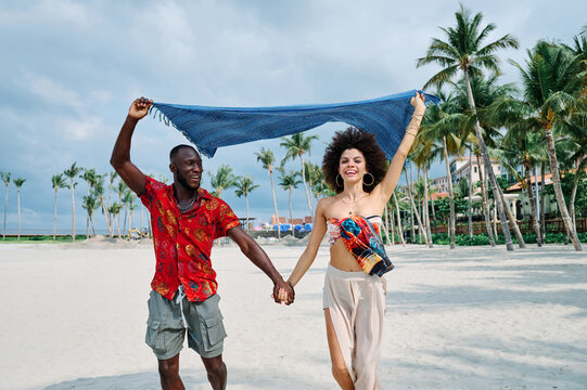 A joyful couple runs hand in hand along a sandy beach near a yacht and modern marina, laughing and waving a blue scarf overhead. Warm tropical palms, bright clothing and carefree movement convey roman