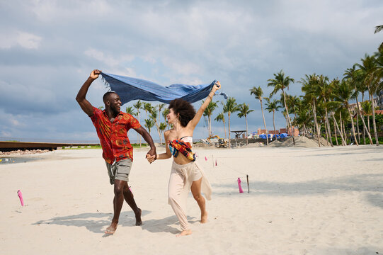 A joyful couple runs hand in hand along a sandy beach near a yacht and modern marina, laughing and waving a blue scarf overhead. Warm tropical palms, bright clothing and carefree movement convey roman