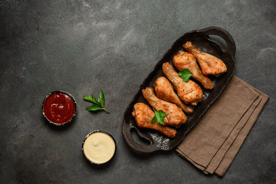 Roasted chicken drumsticks in cast-iron cookware with ketchup and mayonnaise, dark rustic background, top view, flat lay, copy space