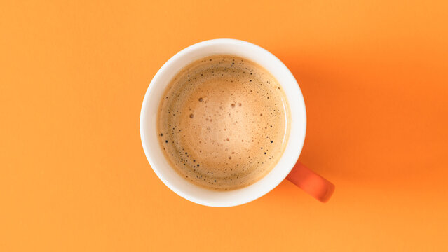 Minimalist autumn background with a single cup of hot coffee with froth on bright orange backdrop. Flat lay top view close up conceptual layout or banner design with empty copy space for text.