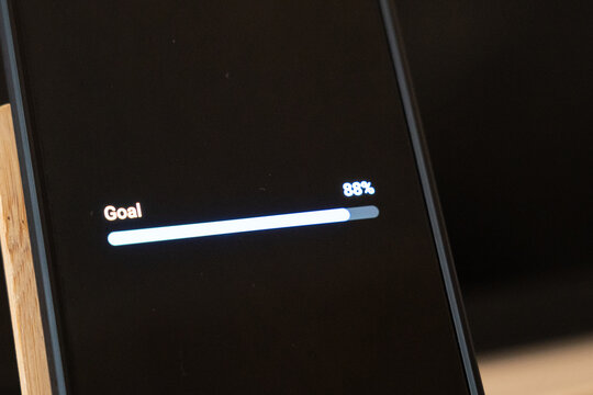 Smartphone screen showing a goal progress bar at 88 percent on a dark interface, phone at an angle on a wooden desk, concept of goal tracking, almost done, productivity, motivation and personal achie