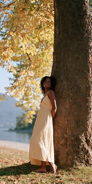 Woman in cream maxi dress leaning against tree on lakeside in golden autumn light for fashion background and lifestyle use