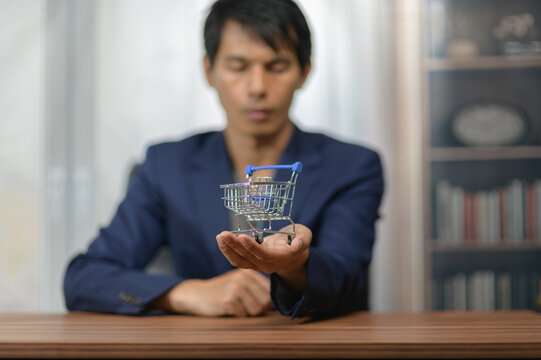 Young Man Holding Miniature Shopping Cart in Focus on Wooden Table