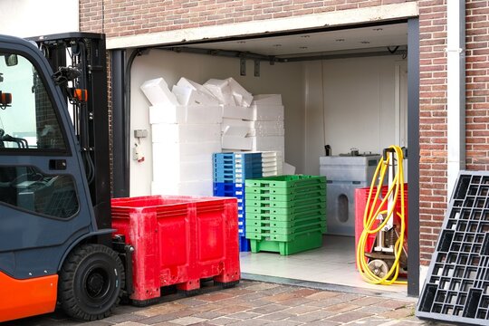 Orange forklift delivering a red plastic bulk container to an open storage room with stacked crates and styrofoam boxes at a fish shop
Forklift carrying a large red plastic container in