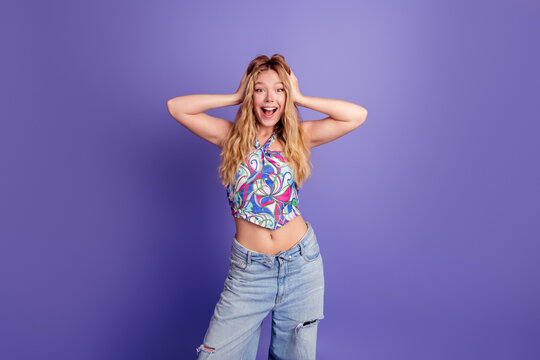 Young woman in colorful halter top and ripped jeans smiling excitedly with hands on head, surprised and joyful portrait against a vibrant purple background for fashion and lifestyle