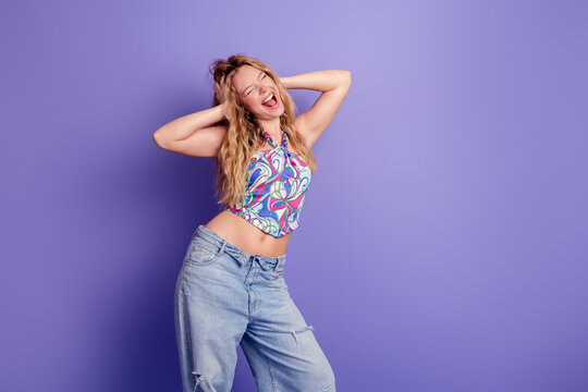 Joyful young woman dancing and singing in colorful retro top and jeans against a purple background, carefree portrait conveying fun and summer fashion lifestyle