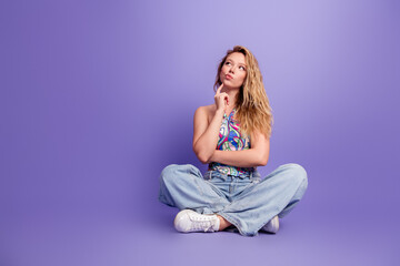 Obraz premium Young woman sitting cross-legged on purple background looking thoughtful and pensive in casual jeans and colorful top