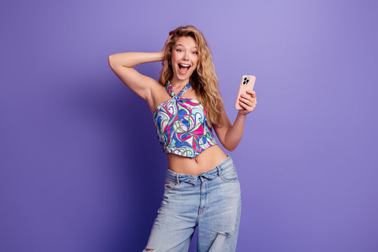 Young woman excitedly holding smartphone against purple background, smiling in colorful summer top and ripped jeans