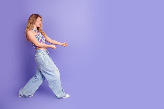 Young woman in wide jeans and colorful crop top playfully miming pulling an invisible rope on a purple studio background, smiling and stepping forward in an energetic casual lifestyle pose
