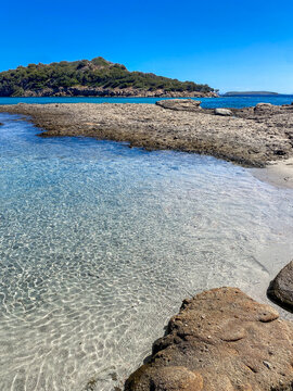 Scenic landscape of secluded Plage de Saint Costanza beach featuring transparent shallow water, foreground rocks, and a view facing the Ile de Farina island near Porto Vecchio, South Corsica, France.