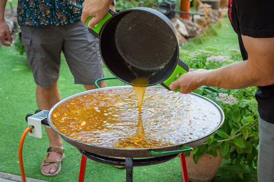 Preparaci&oacute;n de una enorme paella espa&ntilde;ola al aire libre, con caldo caliente verti&eacute;ndose desde una cazuela sobre el arroz en una paellera tradicional