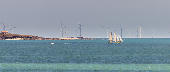 Voilier ancien deux mâts, toutes voiles dehors navigant devant une île et des éoliennes offshore. © YVO-Photos