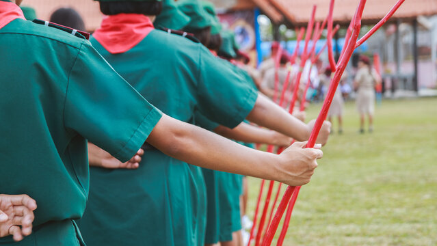 Row of Girl Scouts in Green Uniform Holding Red Wooden Staffs in School Yard