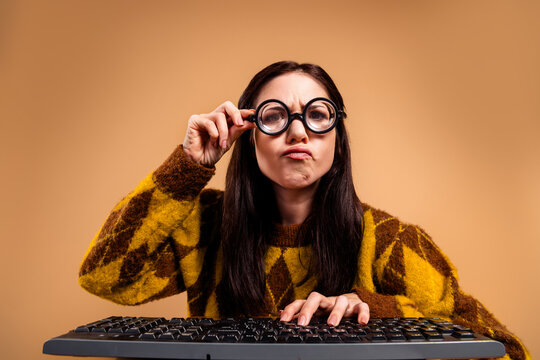 Woman wearing round glasses making a funny confused face while typing on a keyboard in a retro sweater, portrait of a quirky programmer or student at a computer