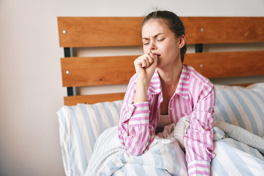 A young woman in pink striped pajamas coughs into her fist while sitting in bed at home. Concept of morning sickness, cold, flu and illness.