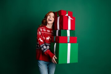 Naklejka premium Happy young woman in red Christmas sweater carrying a tall stack of wrapped gifts against a green background